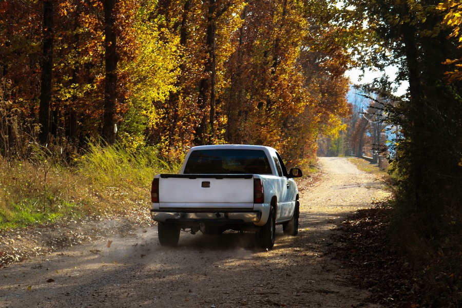 A white pickup truck driving down a dirt road during the daytime. The road is surrounded by tall trees.