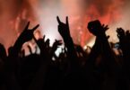 A crowd of concert goers raises their hands in a heavy metal gesture, while a band plays on stage in the background.
