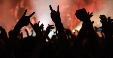 A crowd of concert goers raises their hands in a heavy metal gesture, while a band plays on stage in the background.