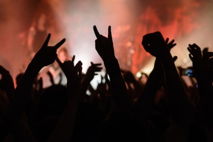 A crowd of concert goers raises their hands in a heavy metal gesture, while a band plays on stage in the background.