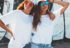 Two women in white oversized tees and colorful round sunglasses pose together outdoors against a graffiti wall.