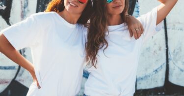Two women in white oversized tees and colorful round sunglasses pose together outdoors against a graffiti wall.