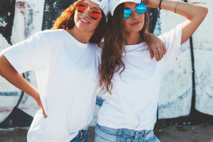 Two women in white oversized tees and colorful round sunglasses pose together outdoors against a graffiti wall.