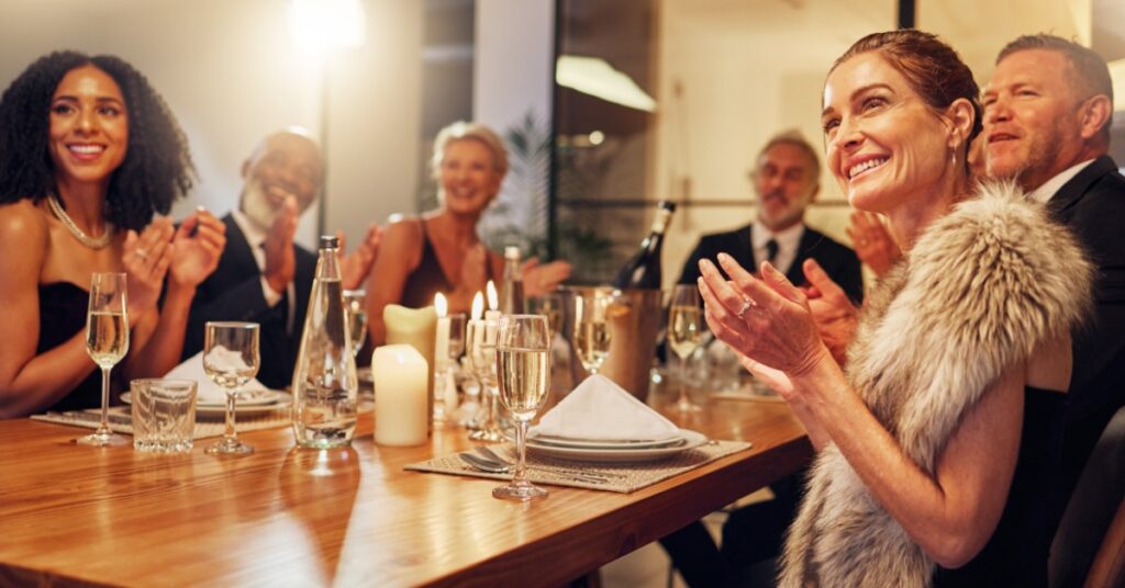 A group of people in formal clothing clapping around a wooden restaurant table with candles, glasses, and plates.