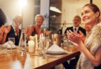 A group of people in formal clothing clapping around a wooden restaurant table with candles, glasses, and plates.