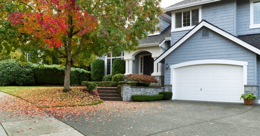 A blue two-story house with a white garage, a curved driveway, front steps, and red leaves scattered under a tree.