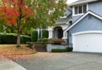 A blue two-story house with a white garage, a curved driveway, front steps, and red leaves scattered under a tree.