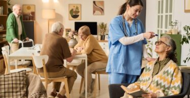 An older woman holding a book as a woman in scrubs checks her temperature, with older adults talking behind them.
