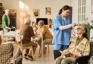 An older woman holding a book as a woman in scrubs checks her temperature, with older adults talking behind them.