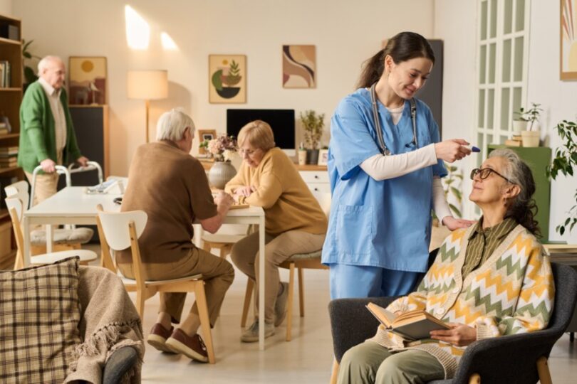 An older woman holding a book as a woman in scrubs checks her temperature, with older adults talking behind them.