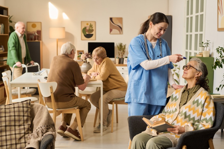 An older woman holding a book as a woman in scrubs checks her temperature, with older adults talking behind them.