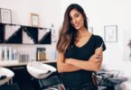 Portrait of a hair stylist holding a comb while standing in a salon with shampoo stations and styling products.