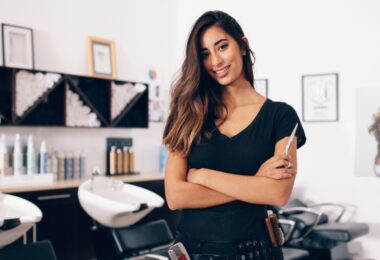 Portrait of a hair stylist holding a comb while standing in a salon with shampoo stations and styling products.