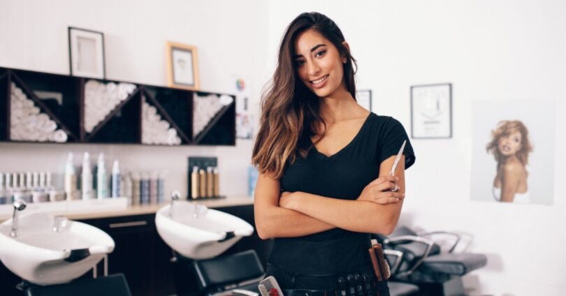 Portrait of a hair stylist holding a comb while standing in a salon with shampoo stations and styling products.