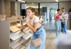 A woman touching a wood flooring sample in a store aisle while two people review materials in the background.
