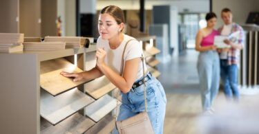 A woman touching a wood flooring sample in a store aisle while two people review materials in the background.