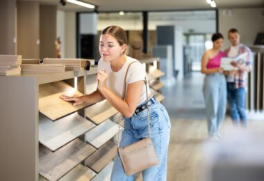A woman touching a wood flooring sample in a store aisle while two people review materials in the background.