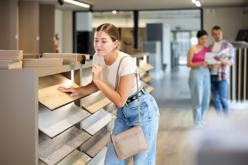 A woman touching a wood flooring sample in a store aisle while two people review materials in the background.