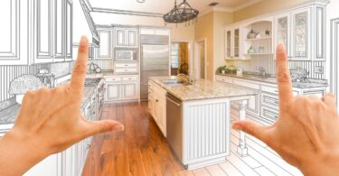 Two hands framing a kitchen design blending a design drawing with finished cabinets and kitchen appliances.