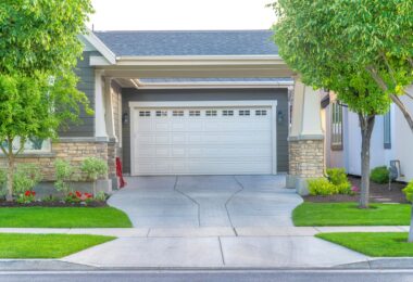 A view from the street of a house's garage with a white door. There is a concrete driveway leading up to the door.