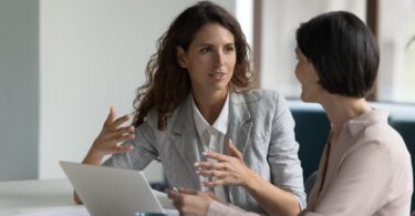 Two women wearing business-casual clothing sit at a table with a laptop open. One woman talks and gestures with her hands.