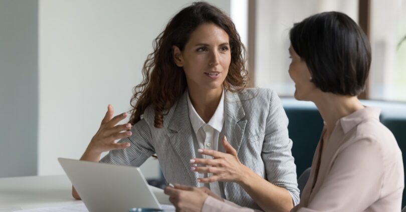 Two women wearing business-casual clothing sit at a table with a laptop open. One woman talks and gestures with her hands.