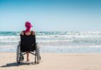 A person with bright pink hair sitting in a wheelchair on a sandy beach facing ocean waves under a blue sky.