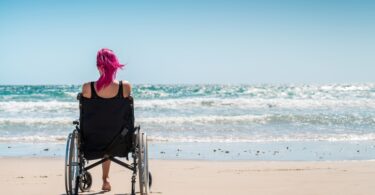 A person with bright pink hair sitting in a wheelchair on a sandy beach facing ocean waves under a blue sky.