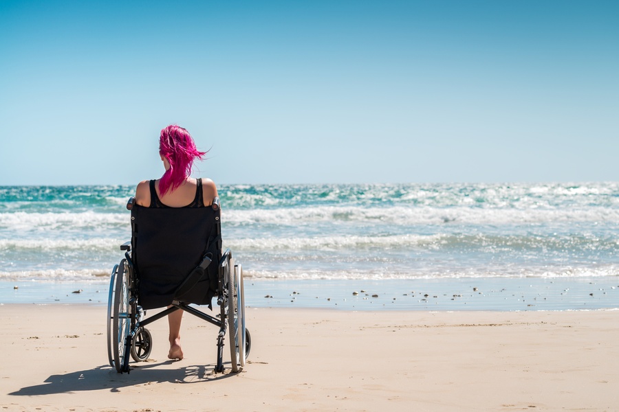 A person with bright pink hair sitting in a wheelchair on a sandy beach facing ocean waves under a blue sky.