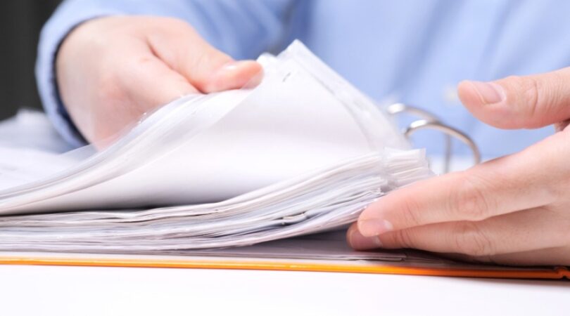 A close-up of a person flipping through a stack of papers in a three-ring binder. The person is wearing a blue shirt.