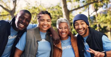 A group of male and female adults in matching blue T-shirts that say "VOLUNTEER" smiling and posing outside together.