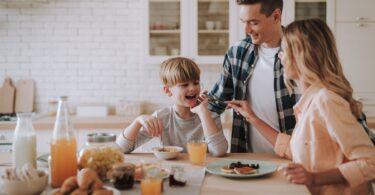 A smiling family gathered around a kitchen table. The young boy eats breakfast with his parents nearby.