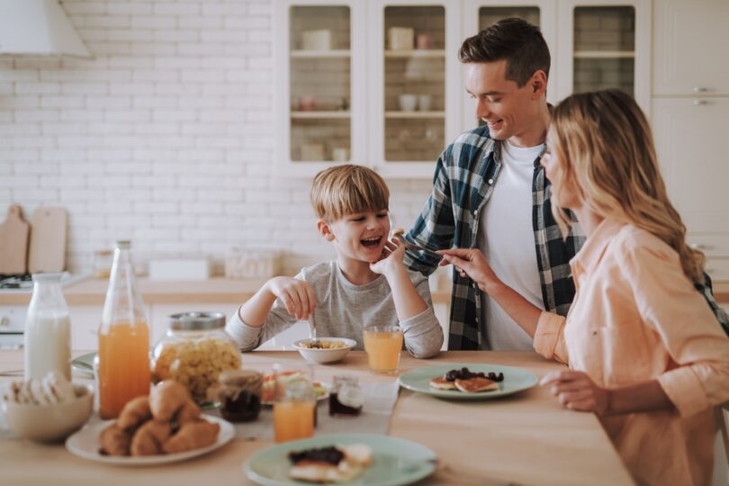A smiling family gathered around a kitchen table. The young boy eats breakfast with his parents nearby.