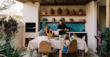 A family of four sitting around a table with a beige tablecloth next to an outdoor kitchen under a wooden roof.