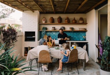 A family of four sitting around a table with a beige tablecloth next to an outdoor kitchen under a wooden roof.