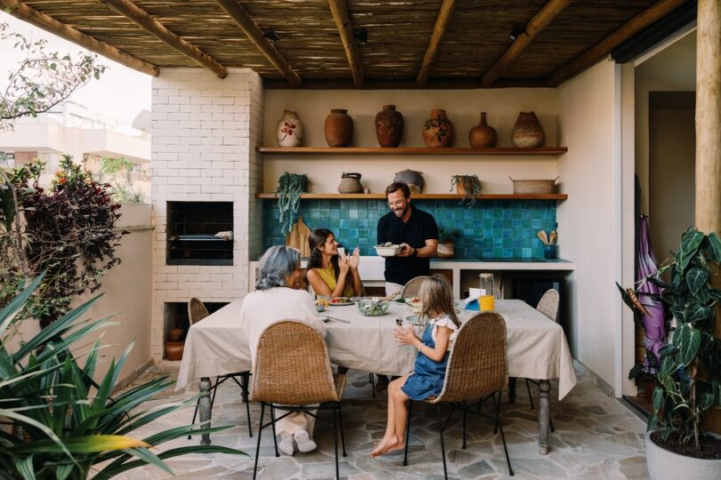 A family of four sitting around a table with a beige tablecloth next to an outdoor kitchen under a wooden roof.
