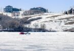 A white pickup truck parked in the middle of a frozen lake with a red tent behind it, with homes in the background.