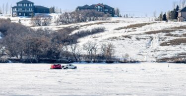 A white pickup truck parked in the middle of a frozen lake with a red tent behind it, with homes in the background.