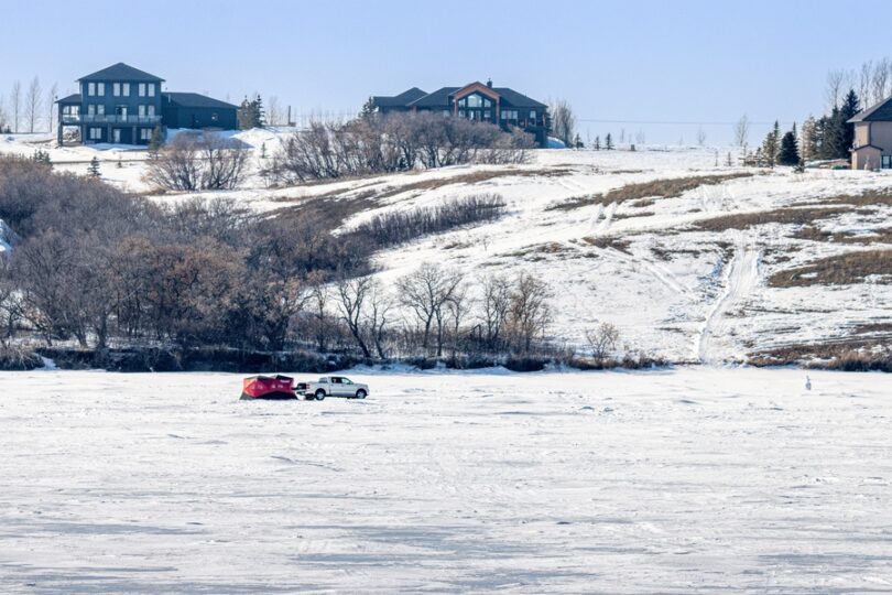 A white pickup truck parked in the middle of a frozen lake with a red tent behind it, with homes in the background.