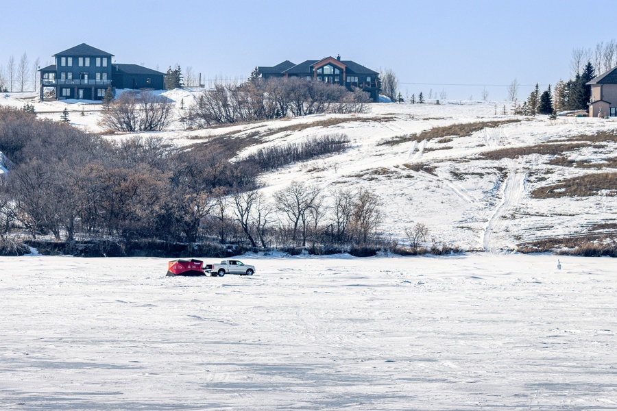 A white pickup truck parked in the middle of a frozen lake with a red tent behind it, with homes in the background.