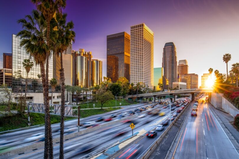 An action shot of the Los Angeles downtown skyline at sunset while cars travel on the nearby highway.