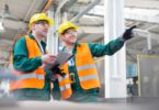 Two workers in yellow hard hats, safety glasses, and orange vests stand in a factory with a clipboard as one points.