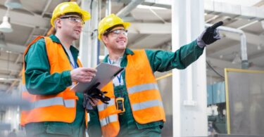 Two workers in yellow hard hats, safety glasses, and orange vests stand in a factory with a clipboard as one points.