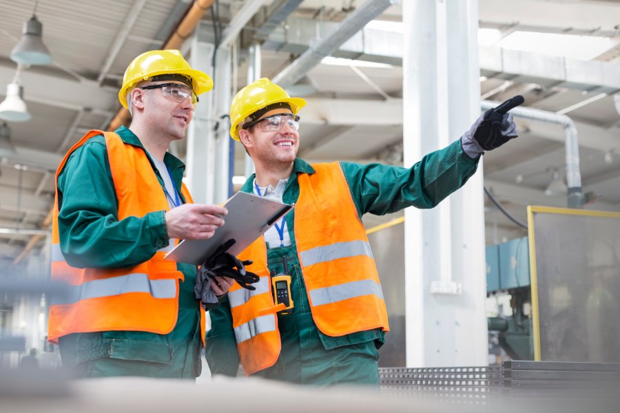 Two workers in yellow hard hats, safety glasses, and orange vests stand in a factory with a clipboard as one points.