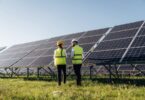 Two people wearing hard hats and yellow safety vests standing in grass beside large rows of solar panels.