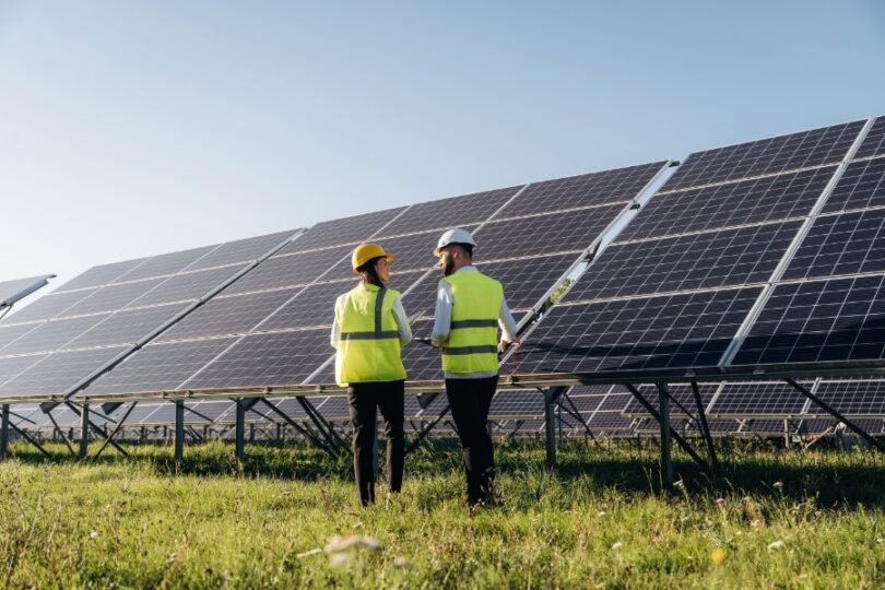 Two people wearing hard hats and yellow safety vests standing in grass beside large rows of solar panels.