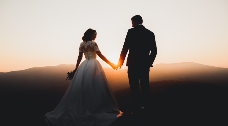 A man in a suit and a woman in a white wedding dress are standing together while they are holding hands.
