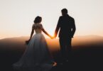 A man in a suit and a woman in a white wedding dress are standing together while they are holding hands.