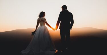 A man in a suit and a woman in a white wedding dress are standing together while they are holding hands.
