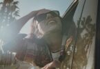 Smiling woman wearing sunglasses leans out of a car window as sunlight filters through a row of palm trees.